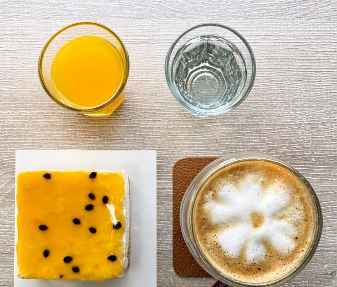 Café con leche con arte latte, cheesecake de maracuyá, jugo y vaso de agua sobre una mesa de madera clara.
